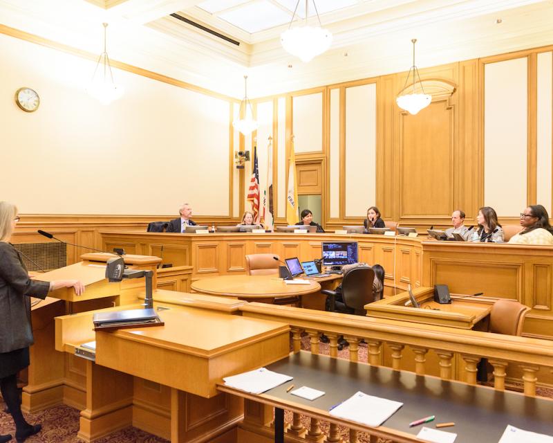 A room with a podium facing a group of people behind a semi-circular table that has SFMTA Board Members speaking with eachother.